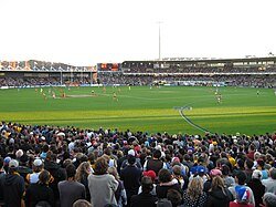 A small stand to the left and a two tier stand and scoreboard filled with people in the backdrop of an oval grass playing surface scattered with players. Spectators stand in the foreground.